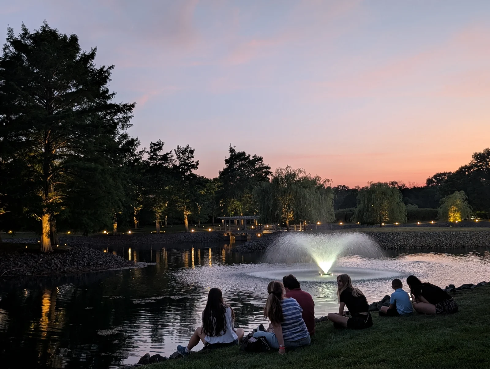 People sitting on the grass beside Zen Lake and its lit fountain at sunset, waiting for fireworks at Bell Works