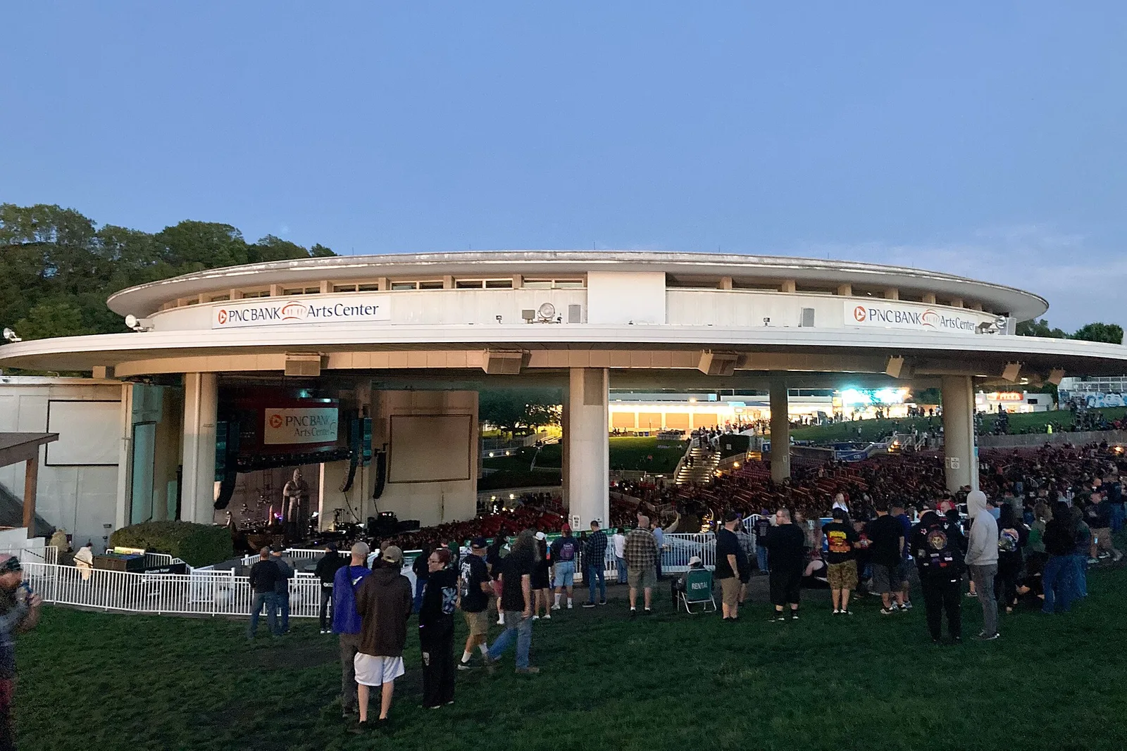 The circular PNC Bank Arts Center amphitheater in Holmdel with a crowd on the lawn at dusk
