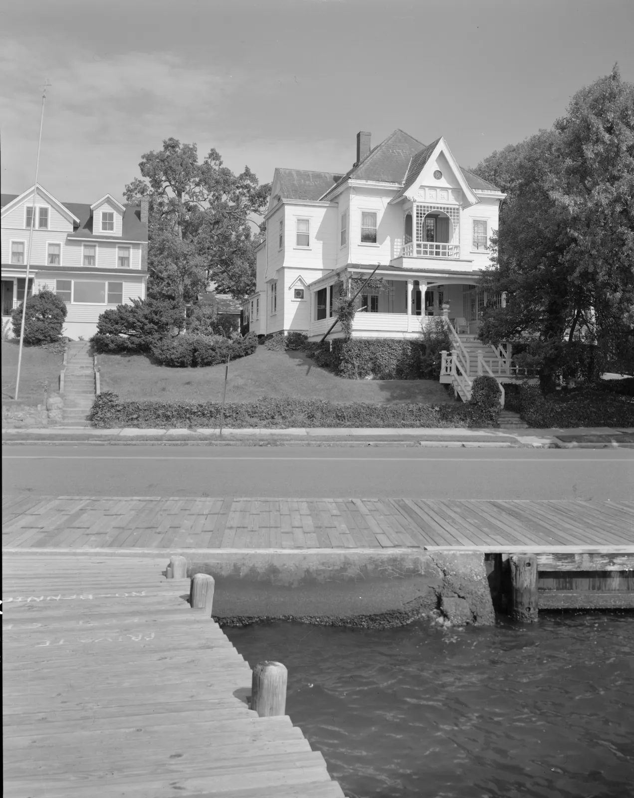 Black-and-white photo of a large Victorian house with a gabled porch on a hill above a riverside dock