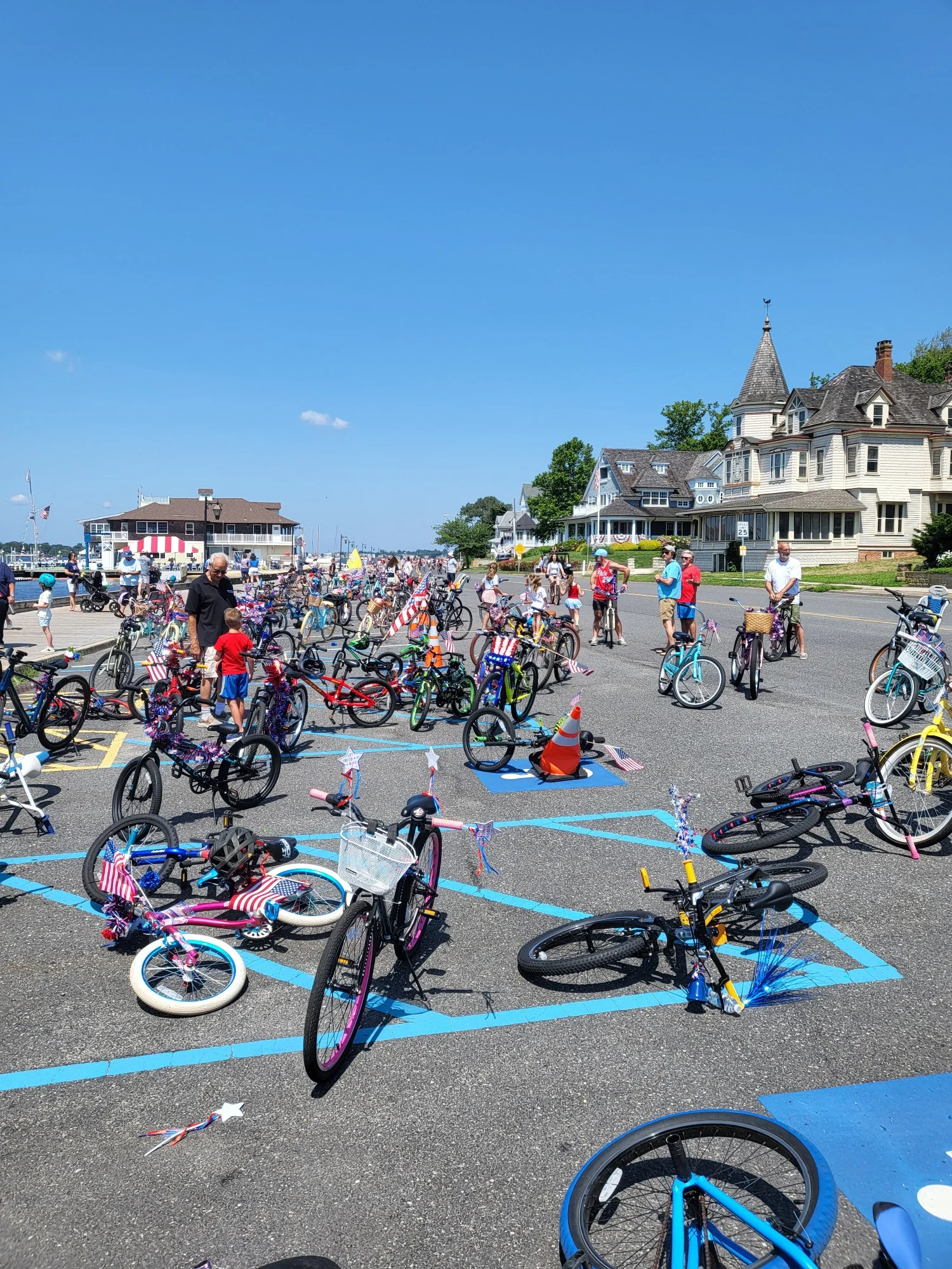 Rows of decorated bicycles lined up near the riverfront for the bike parade, a Victorian home with a turret behind