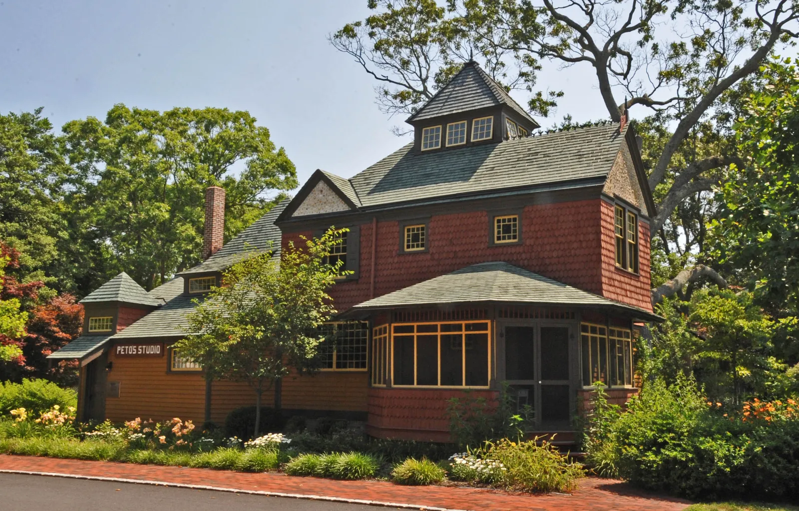 A red shingled Victorian house and studio with a corner bay of windows, surrounded by summer flower beds and shade trees
