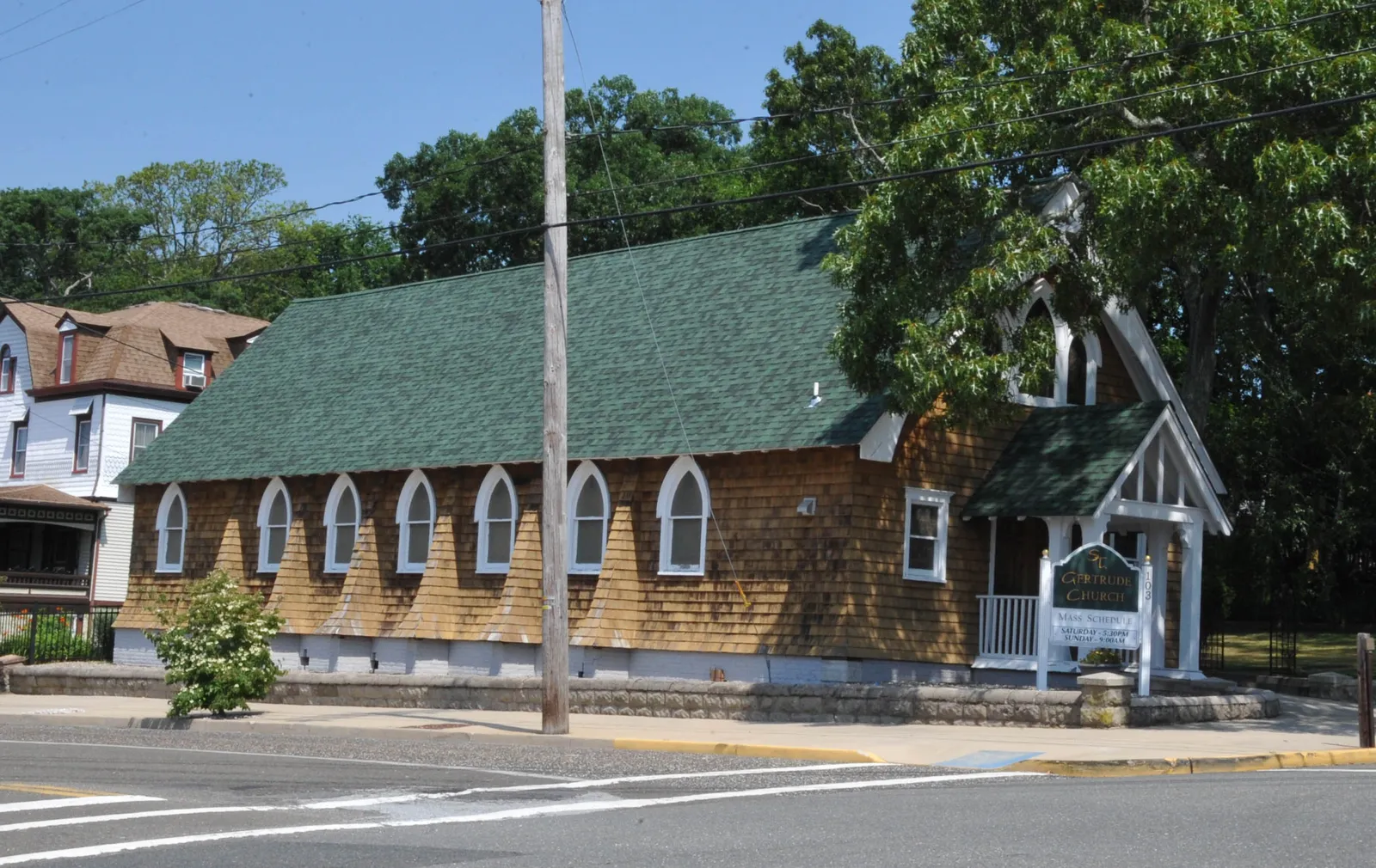A small brown-shingled chapel with pointed white windows and a green roof beside a tree-lined street