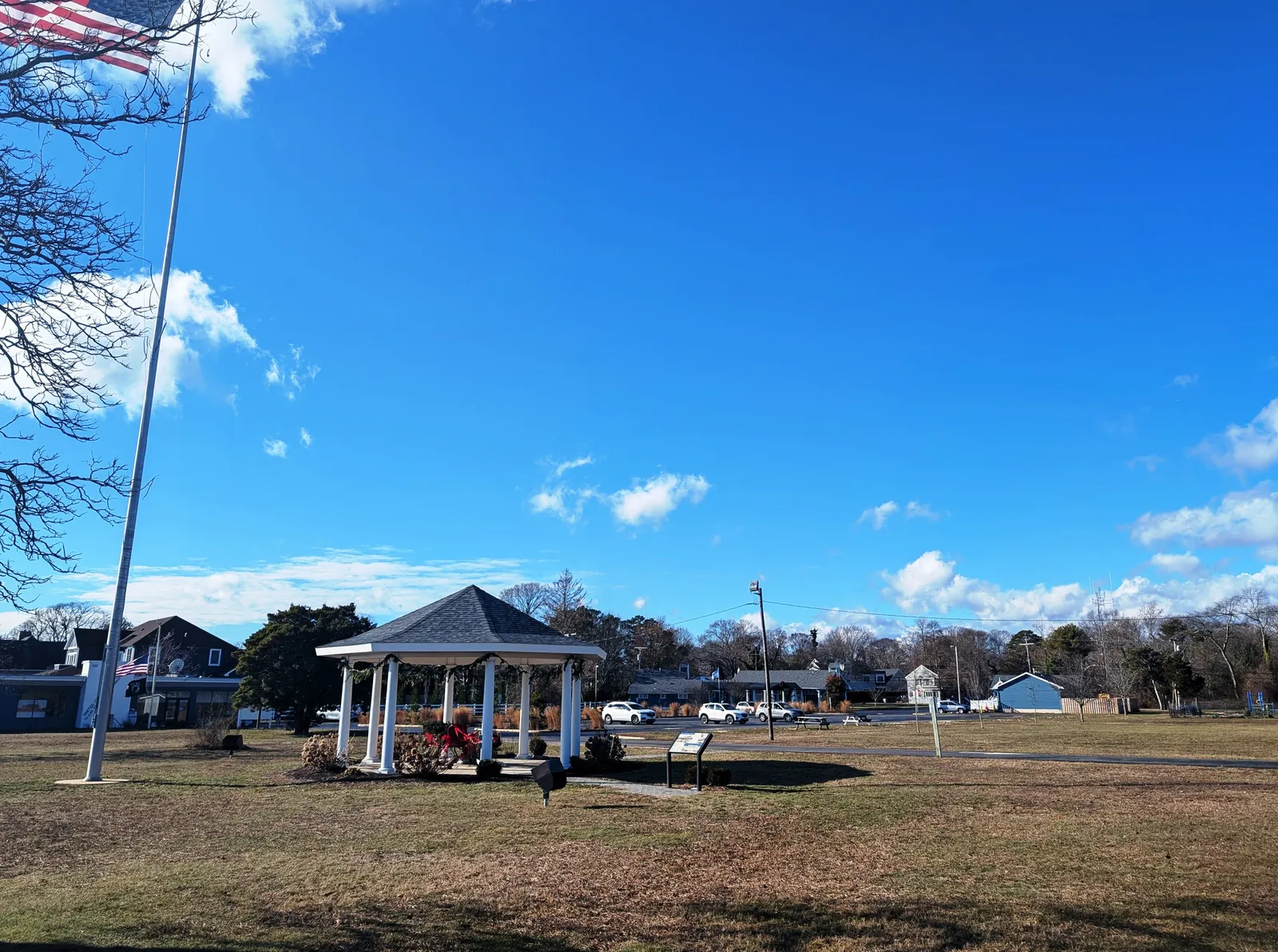 A white gazebo decorated for the holidays on a wide lawn with a tall flagpole under a bright blue sky