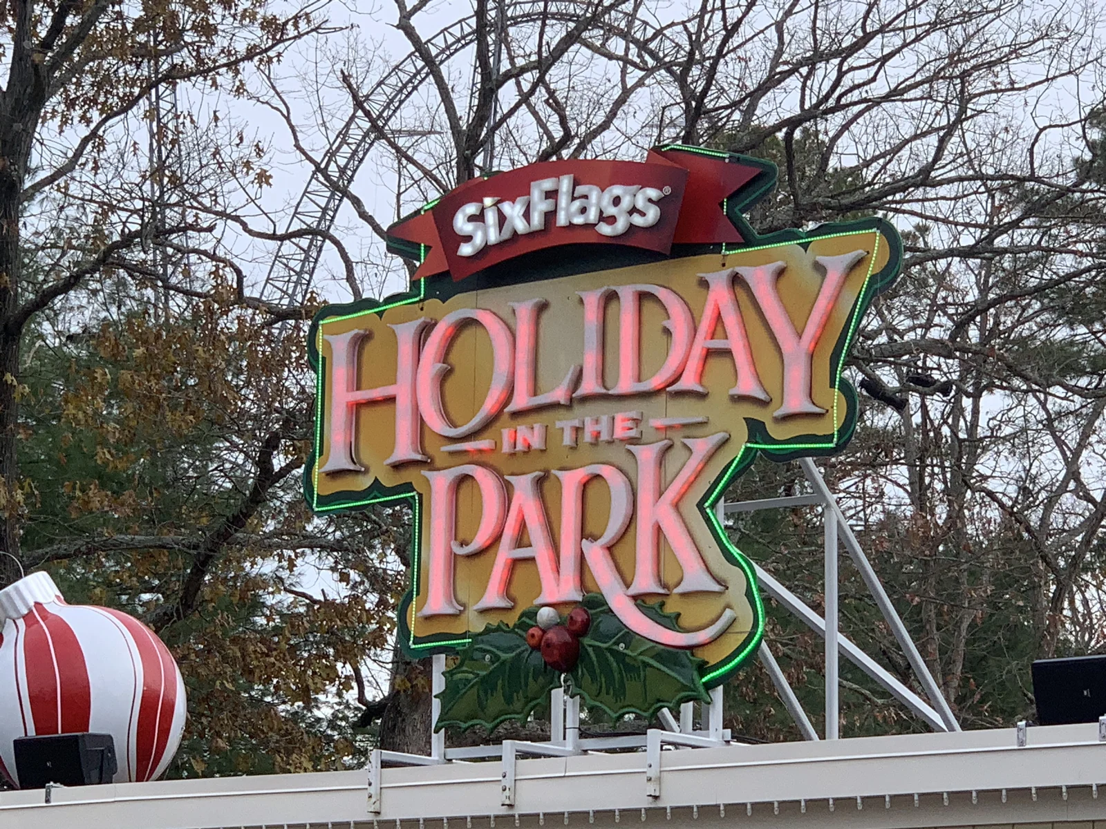 Lit Holiday in the Park sign with holly and ornaments hanging among autumn trees at Six Flags Great Adventure