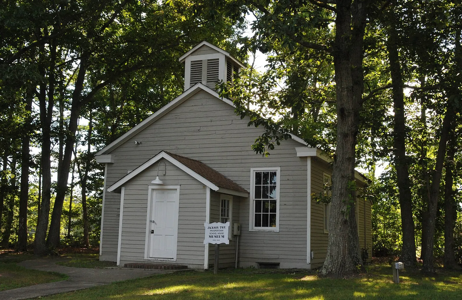 Small gray one-room schoolhouse with a white bell cupola among tall trees