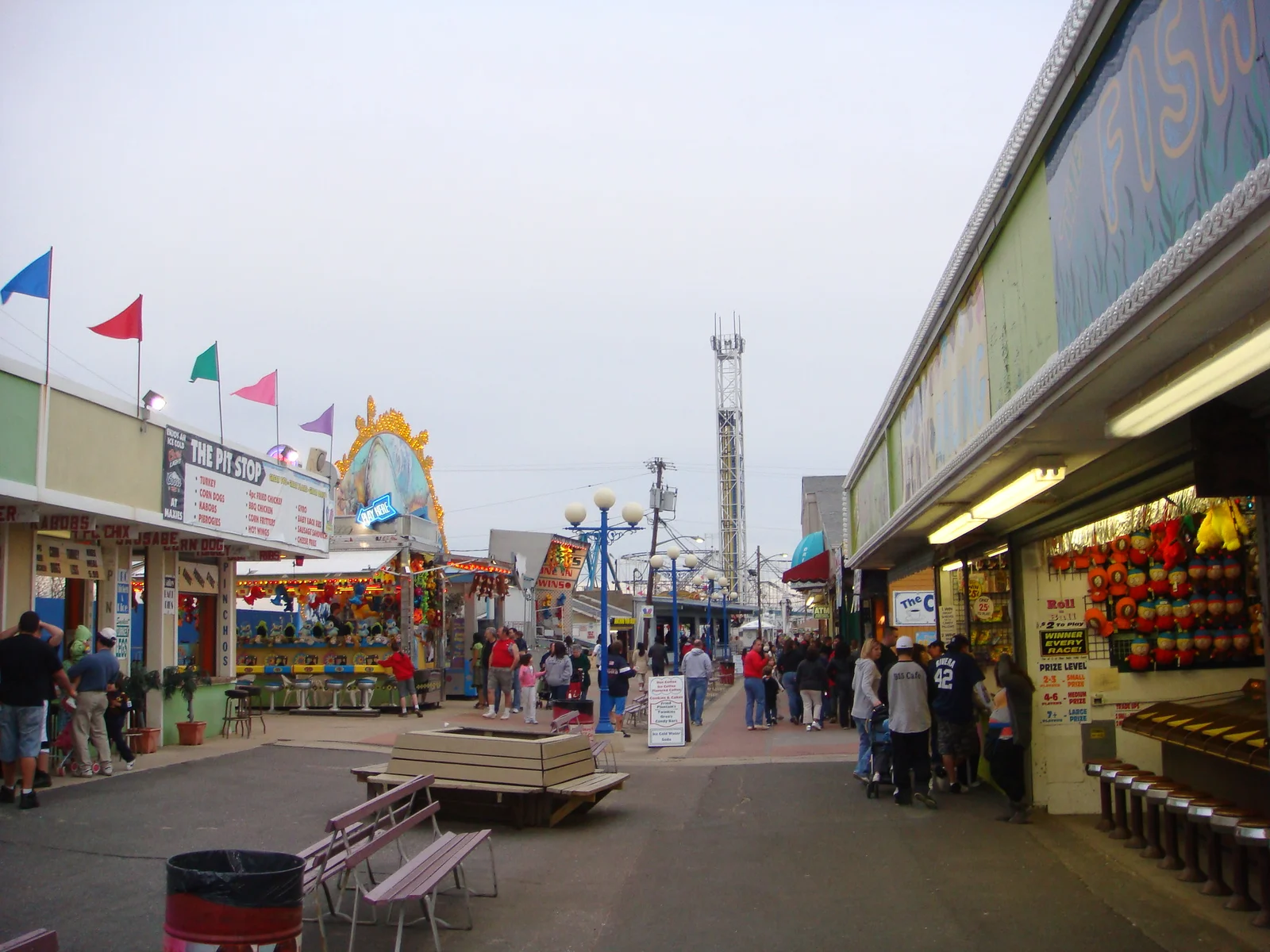 Visitors walking the amusement park midway past game stands, benches and colorful pennant flags, a drop tower in the distance