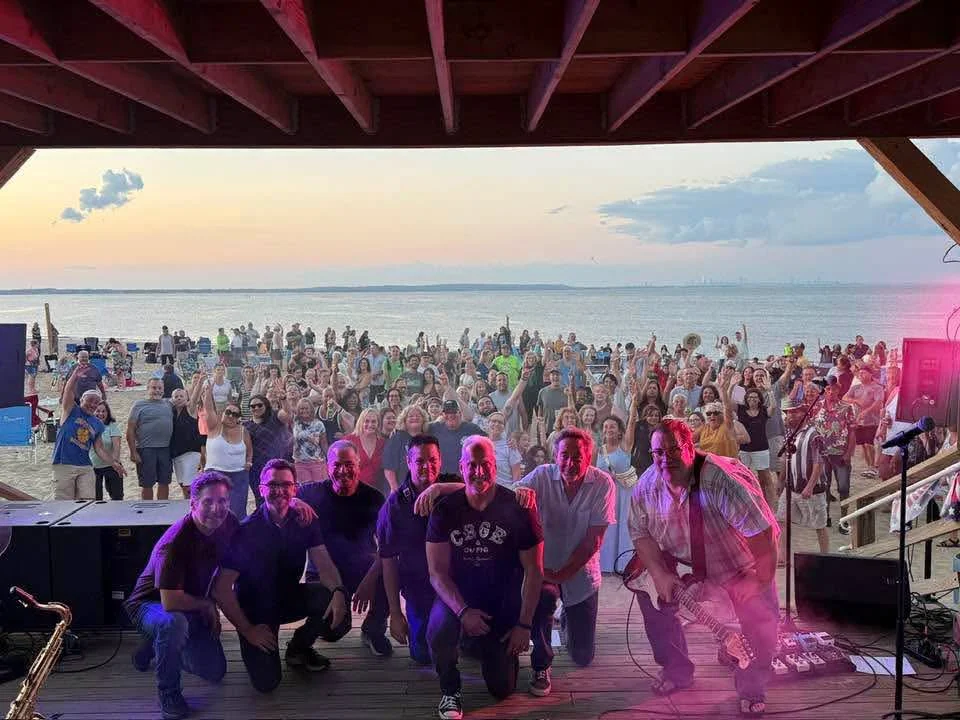 A large crowd on the Keansburg beach cheering toward the stage at sunset, with the band posing at the front of the stage and Raritan Bay behind