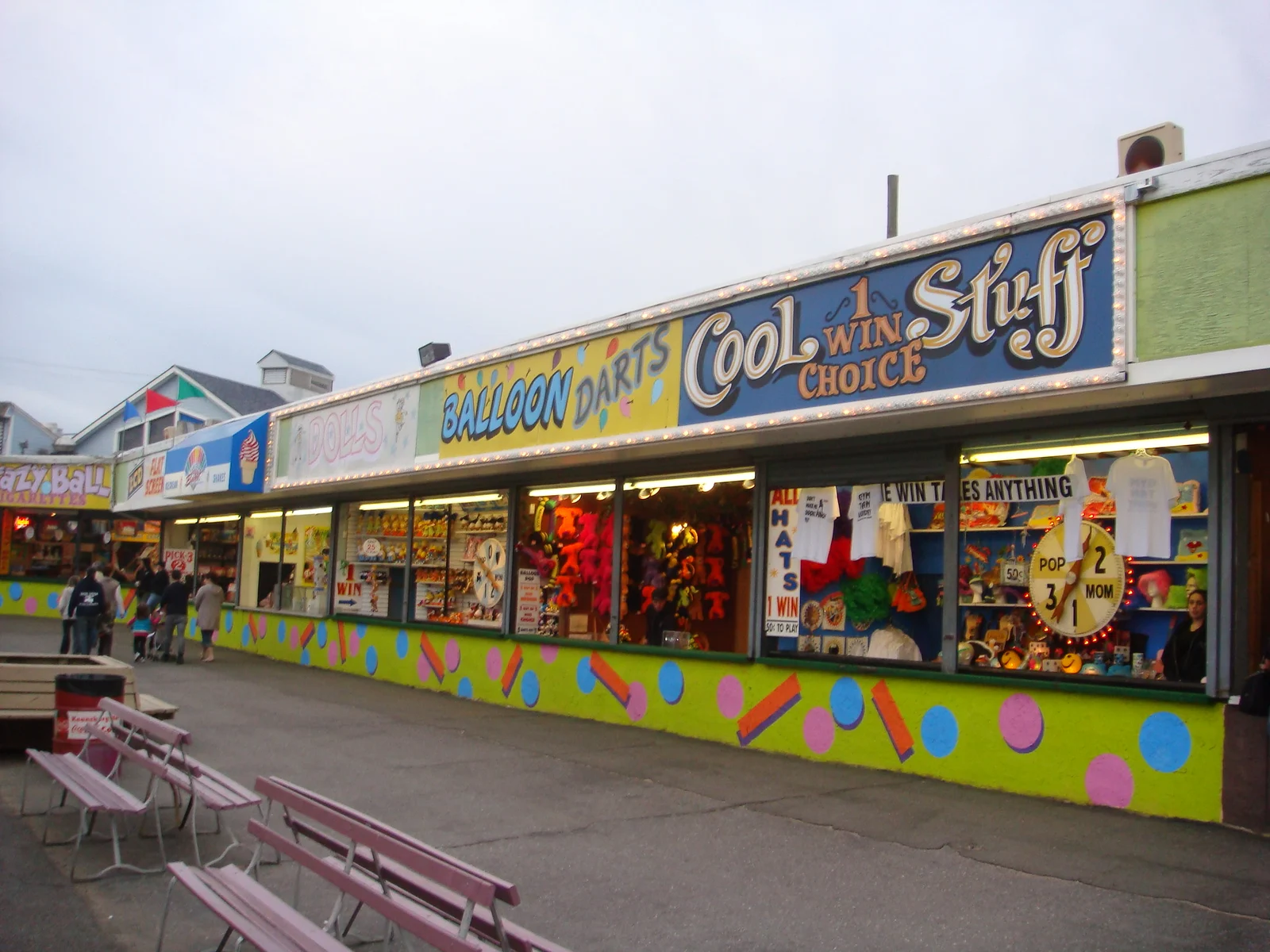 A long row of colorful game booths with prizes, including Balloon Darts and Cool Stuff, along the amusement park walkway