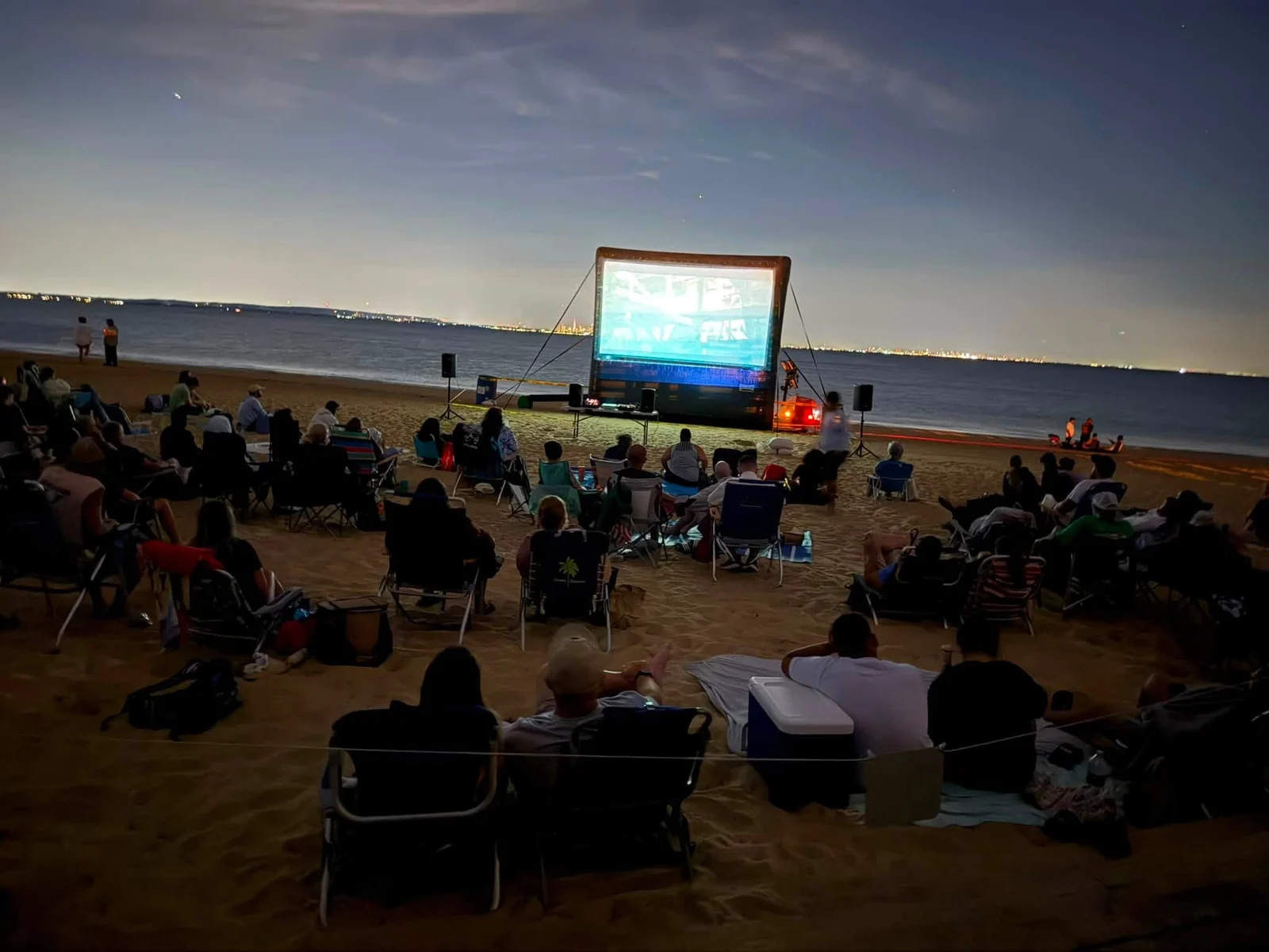 Families in beach chairs on the sand at dusk watching a movie on an inflatable screen, with the lights of the far shore across Raritan Bay