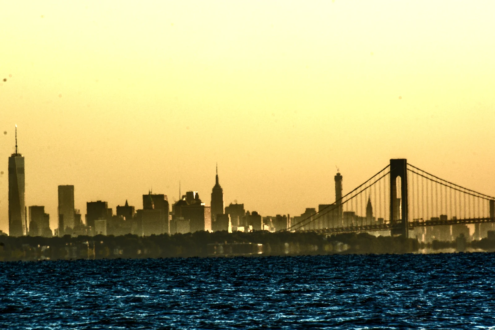 The New York City skyline and the Verrazzano-Narrows Bridge silhouetted against a golden sky across the bay