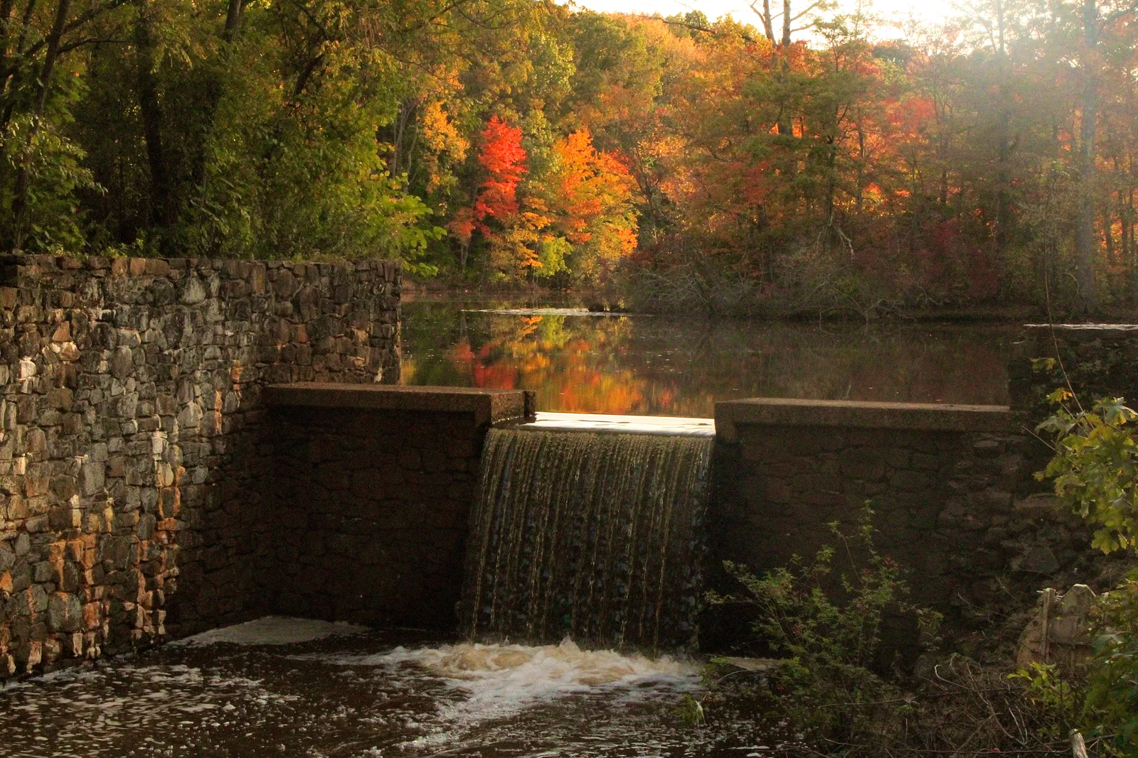 Water spilling over a stone dam below Davidson's Mill Pond, with red, orange and gold fall trees reflected in the still pond