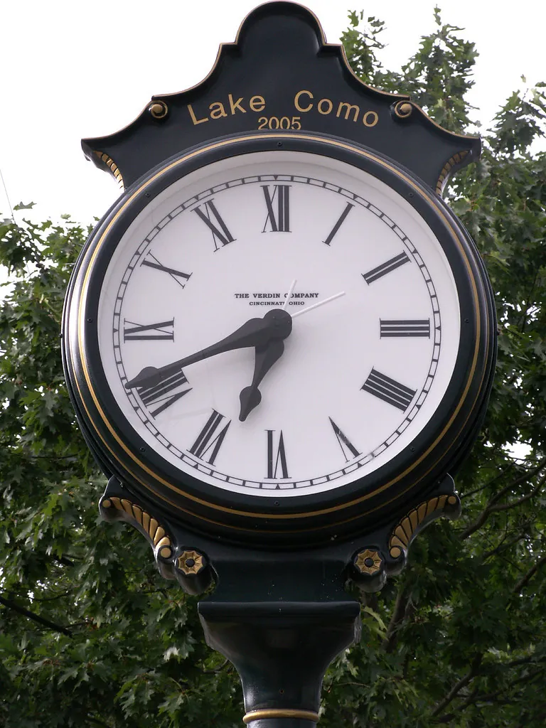 Black post clock with a white face reading Lake Como, against green trees