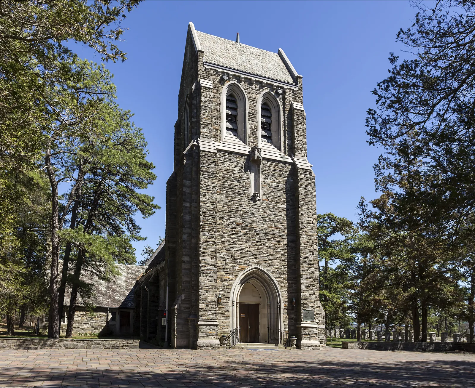 A Gothic stone chapel tower with an arched doorway, framed by pine trees under a clear blue sky
