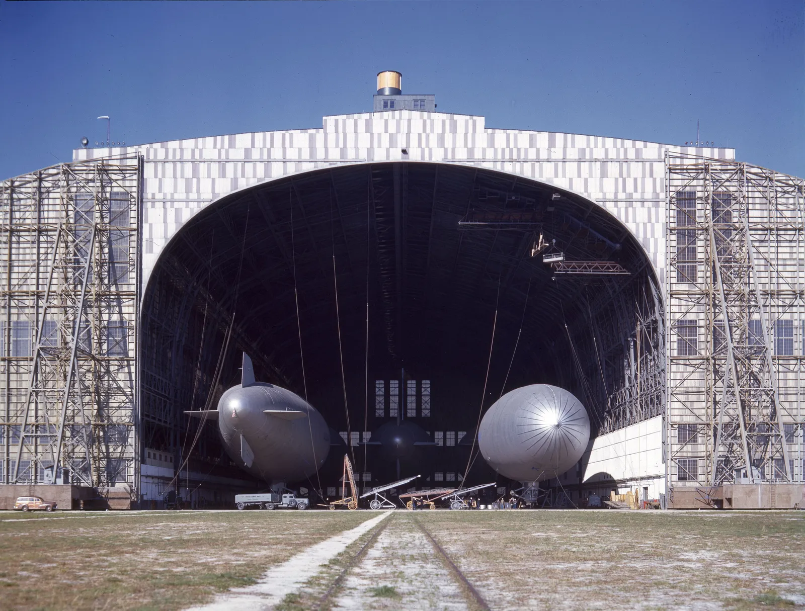 Two silver blimps parked inside the open doors of a giant airship hangar under a blue sky