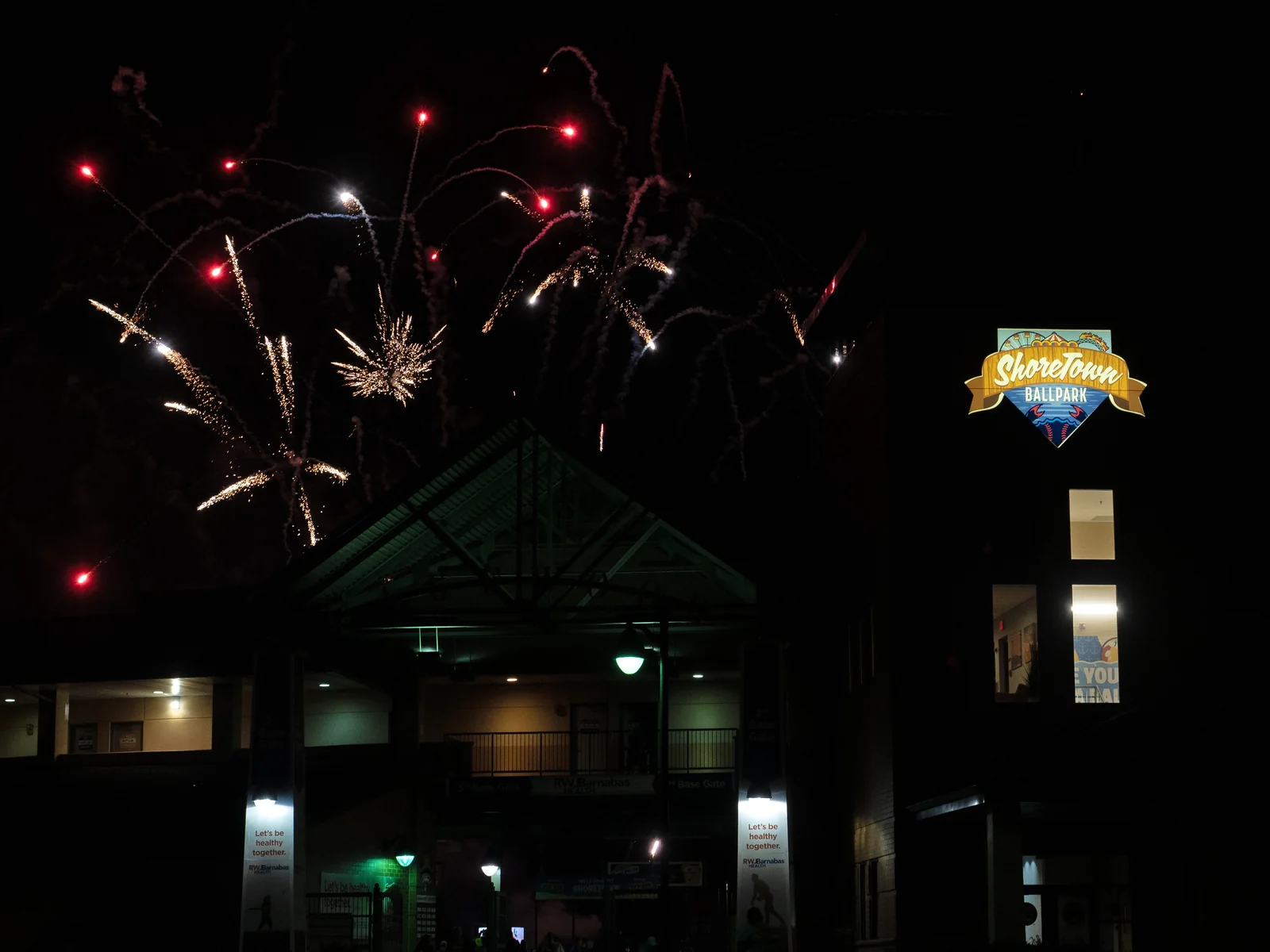 Red and gold fireworks bursting in the night sky above the lit ShoreTown Ballpark entrance and sign