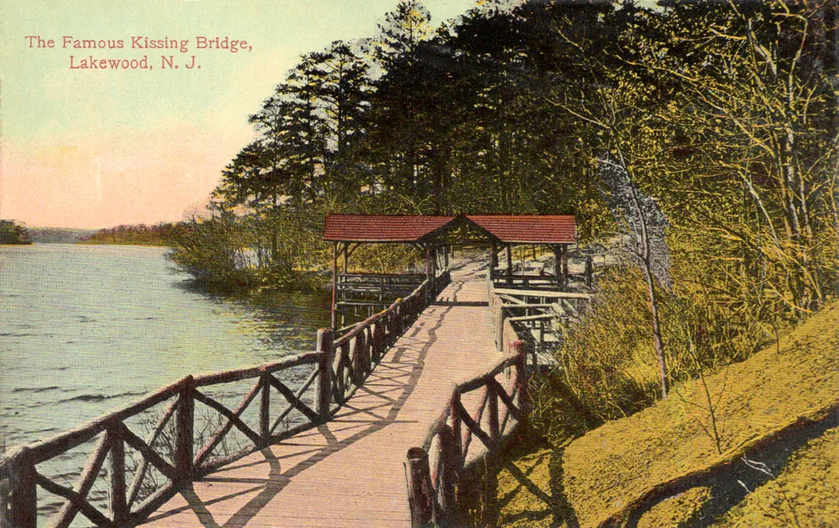 Early color postcard of a wooden footbridge with a small roofed shelter beside the water, framed by trees