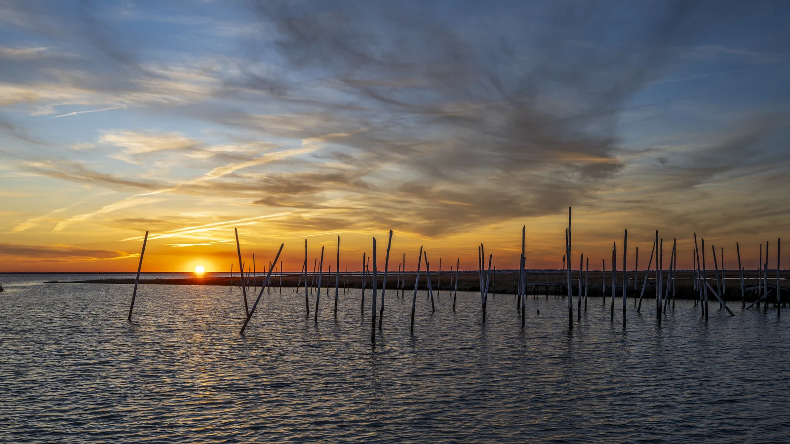 Orange sunset over calm bay water near Great Bay Boulevard, with rows of old wooden stakes standing in the water