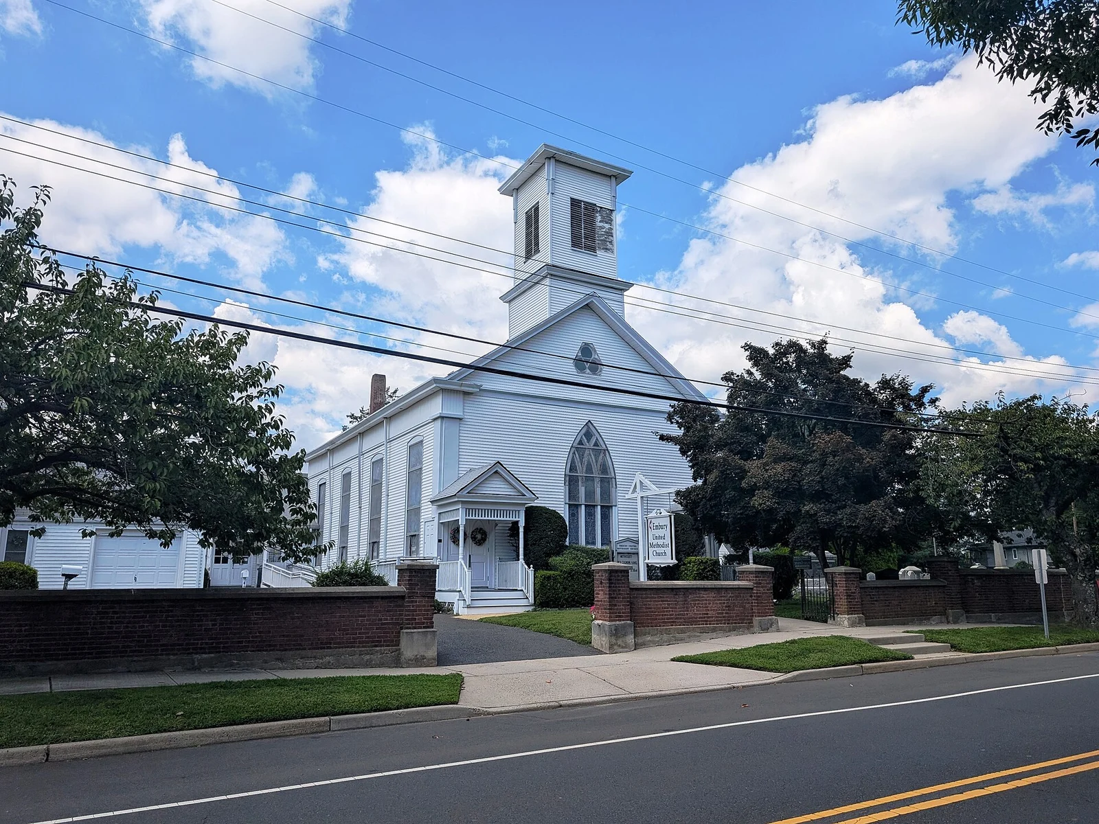 White clapboard Embury United Methodist Church with a square bell tower and pointed-arch windows behind a low brick wall