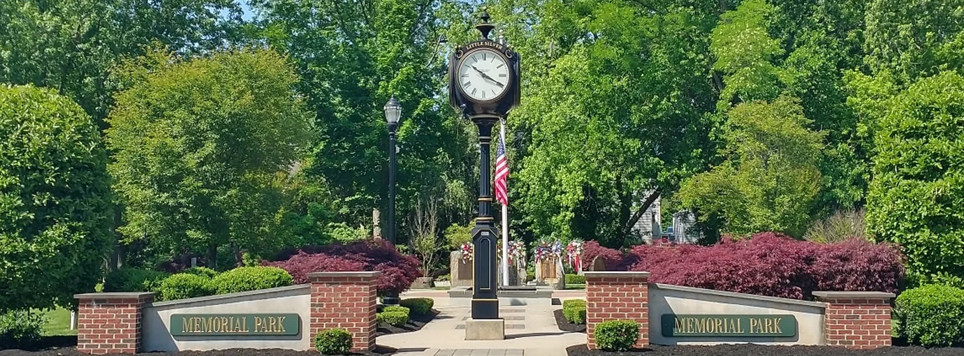 Black post clock and flag at the center of Memorial Park, framed by brick sign walls and red-leafed shrubs