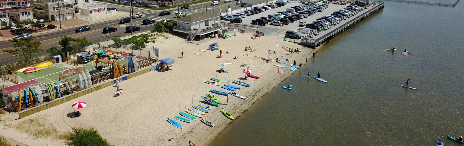 Aerial view of a sandy bay beach lined with rows of paddleboards and kayaks, with paddlers on the calm bay and a board rack beside the beach