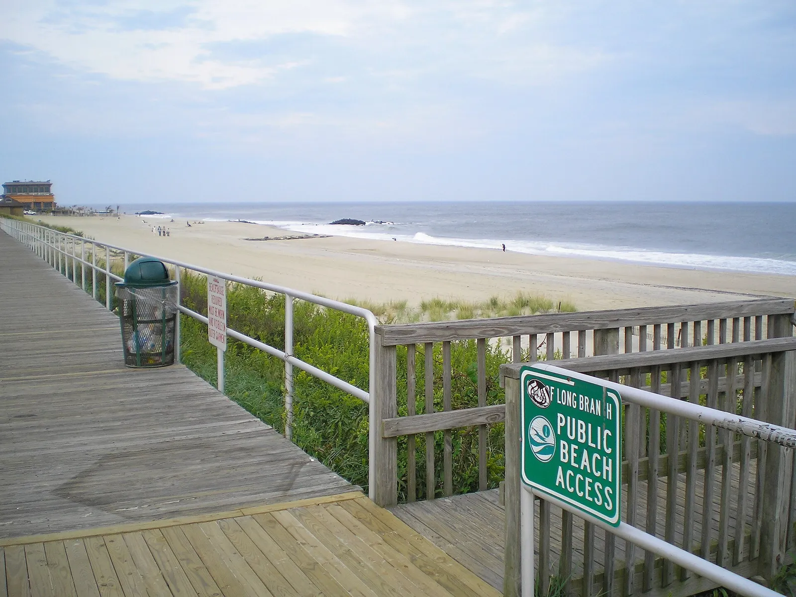 Wooden boardwalk with a railing and a Long Branch public beach access sign beside a wide sandy beach and the ocean