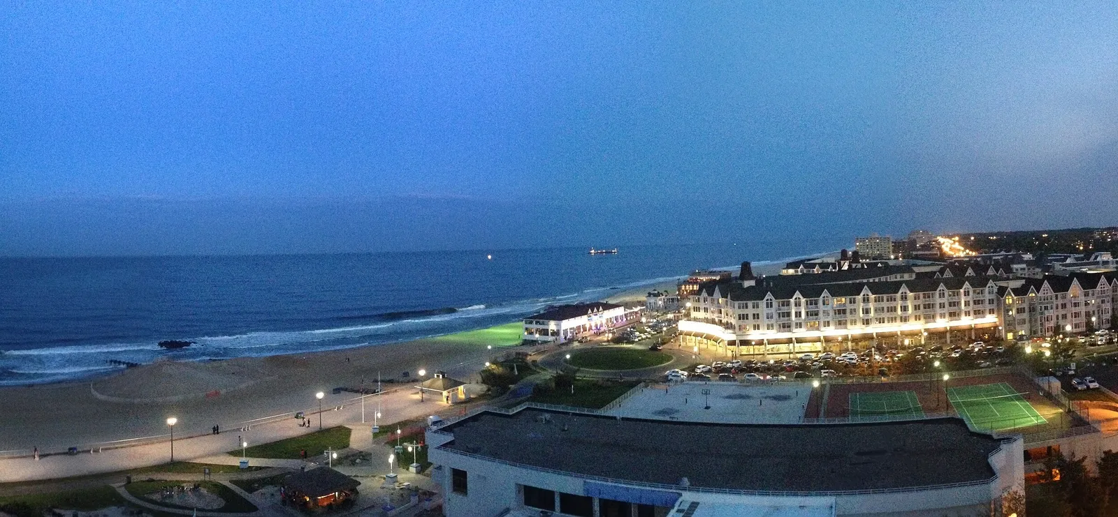 Evening view from above of a lit-up beachfront hotel and shops beside the beach and a boardwalk