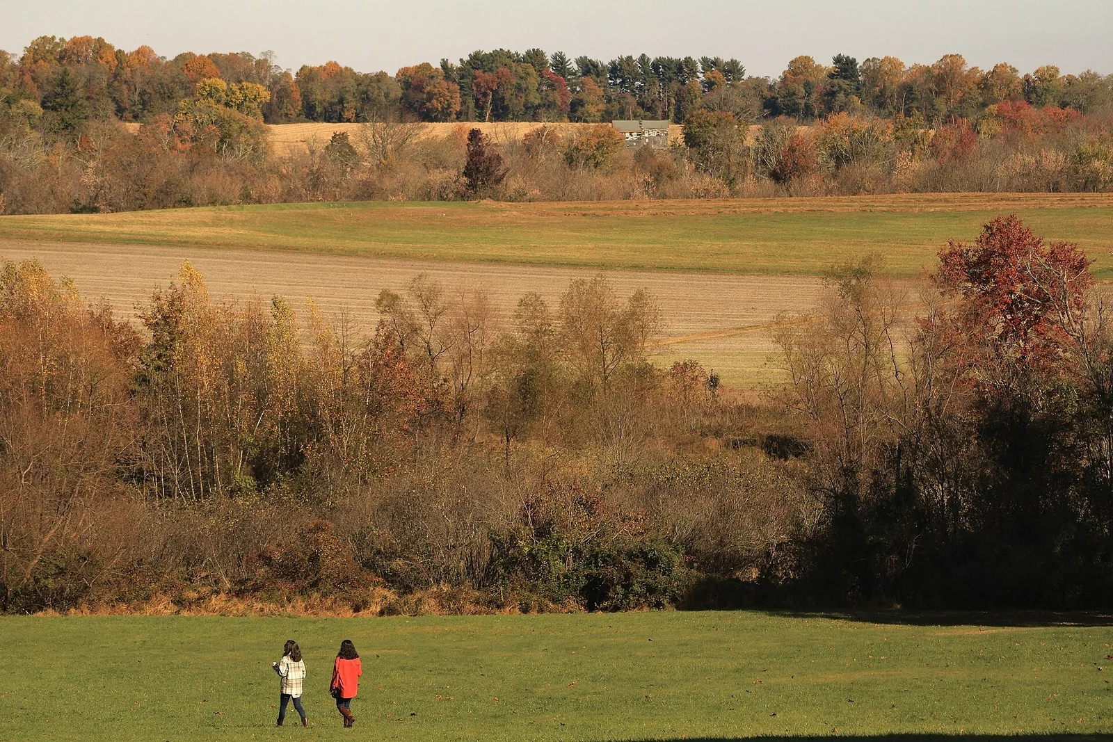 Two walkers crossing a mowed meadow below autumn trees and rolling farm fields at Monmouth Battlefield