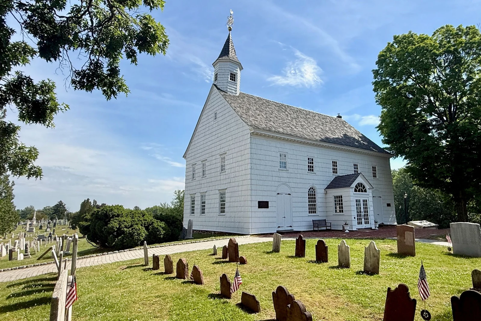 White clapboard Old Tennent Church with its bell tower above rows of old gravestones and small flags