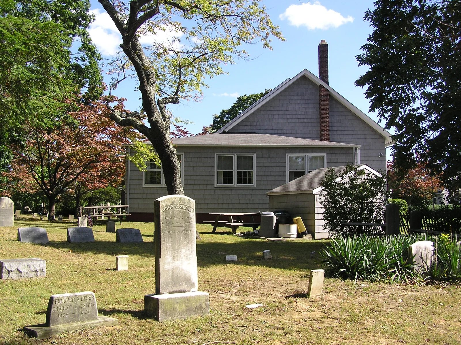 Gray shingled Manasquan Friends Meetinghouse behind old headstones in its burying ground under shade trees