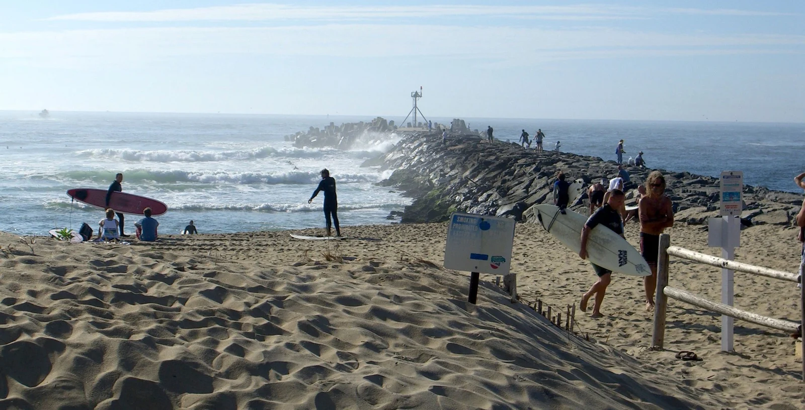 Surf students carrying boards across the sand toward the waves beside the Manasquan Inlet jetty on a sunny morning
