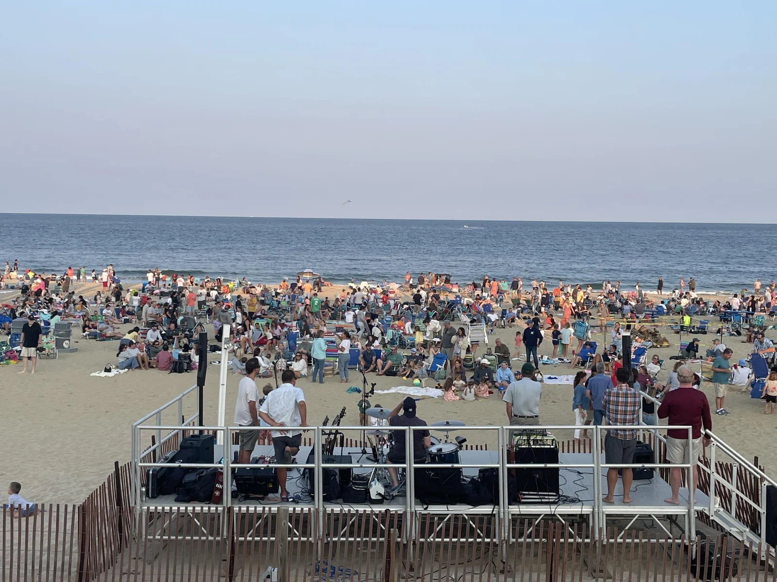 A band on a stage at the edge of Main Beach playing to a large crowd sitting in beach chairs on the sand at sunset