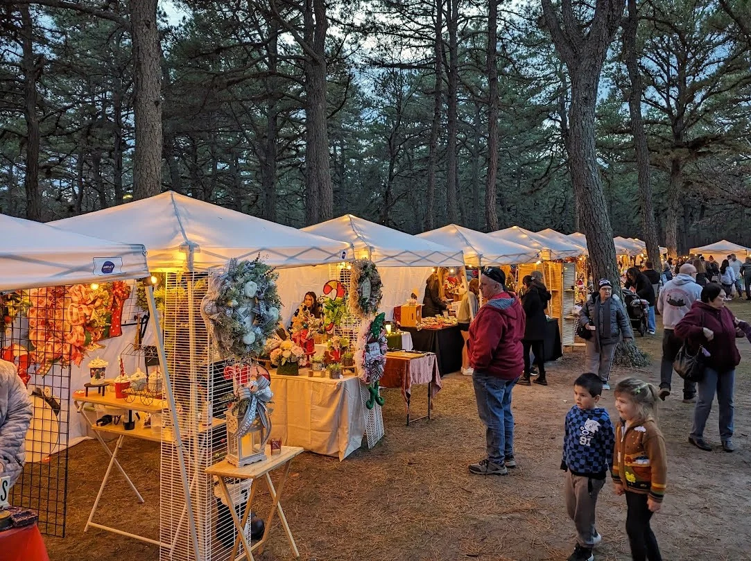 Lit holiday vendor tents with wreaths and gifts under the pines at dusk