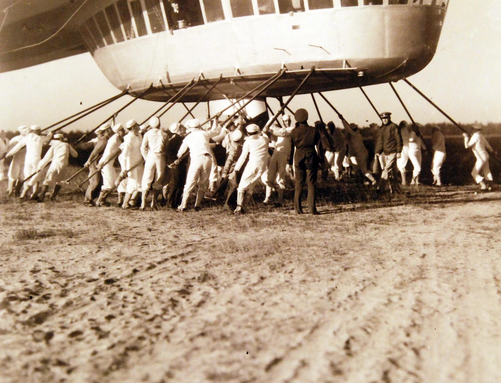 Sepia 1936 photo of a Navy ground crew in white hauling on lines beneath the hull of the airship Hindenburg at Lakehurst