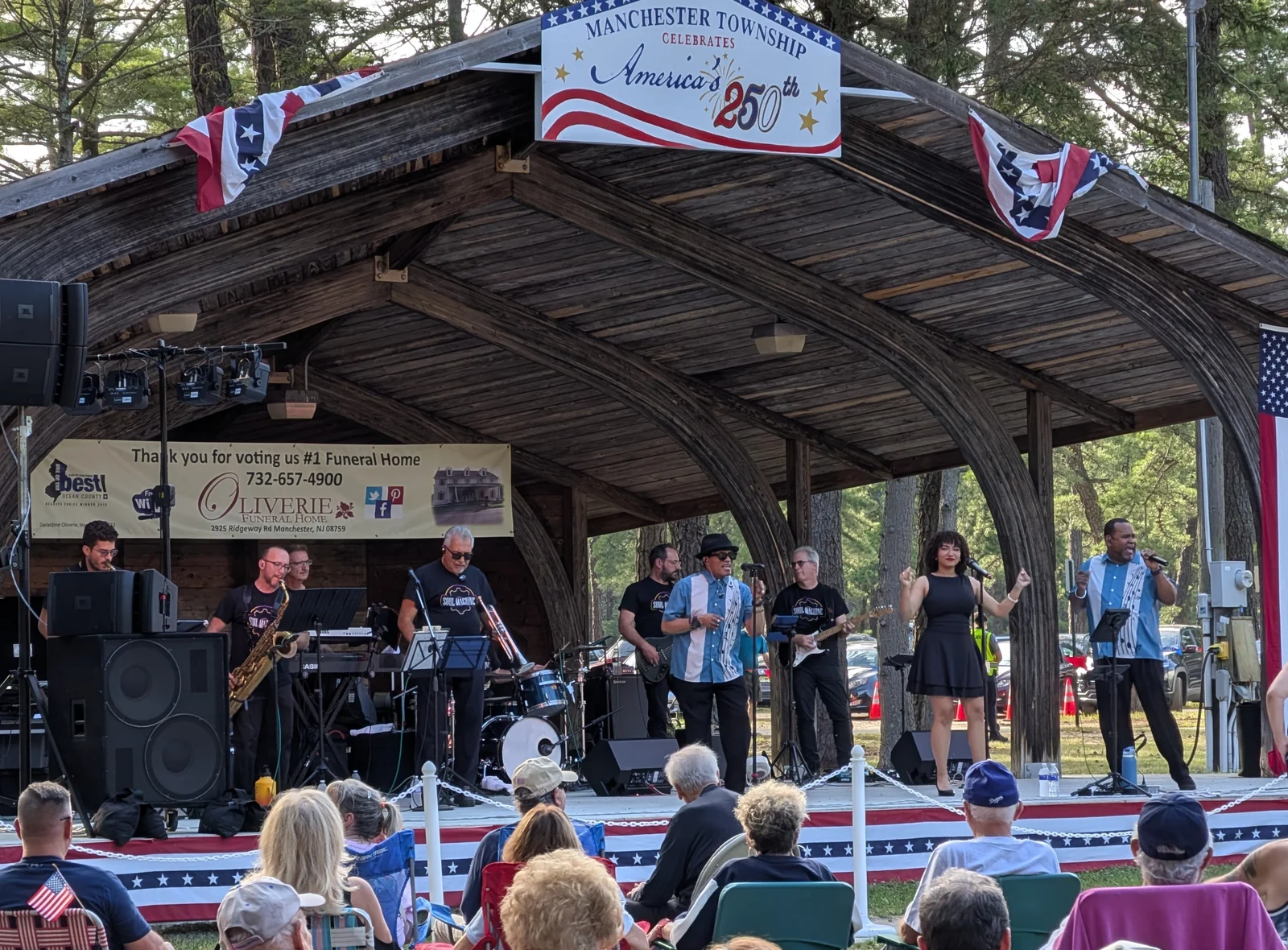 An audience in lawn chairs watches a band under the wooden bandshell at Harry Wright Lake