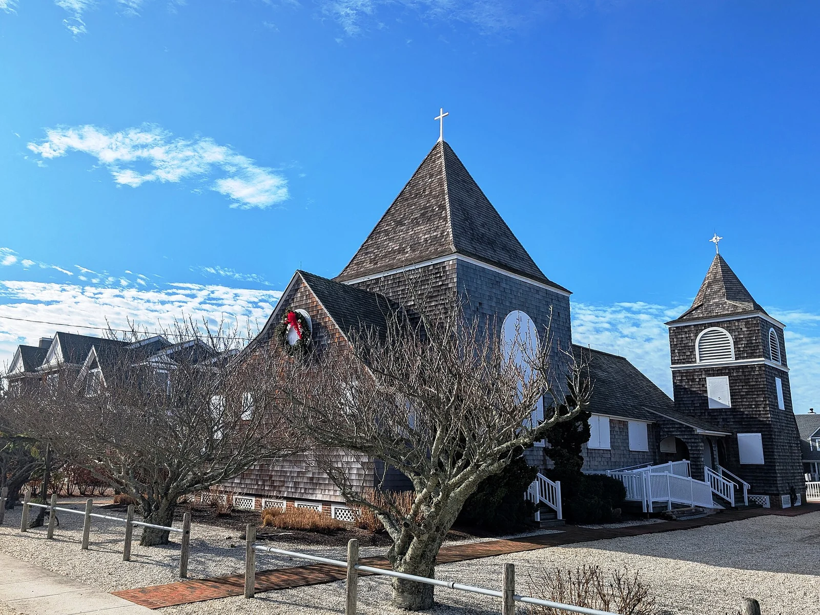 The dark shingled St. Simon By-The-Sea church with its steep peaked roof, bell tower and a wreath over the round window, under a blue sky