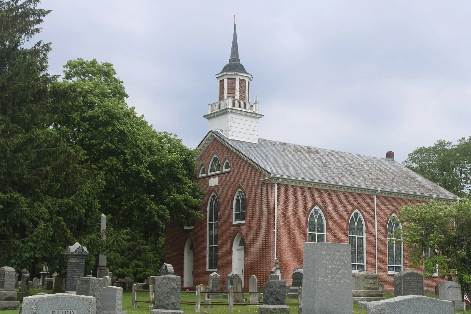 Red brick church with tall arched windows and a white steeple, behind rows of old gravestones