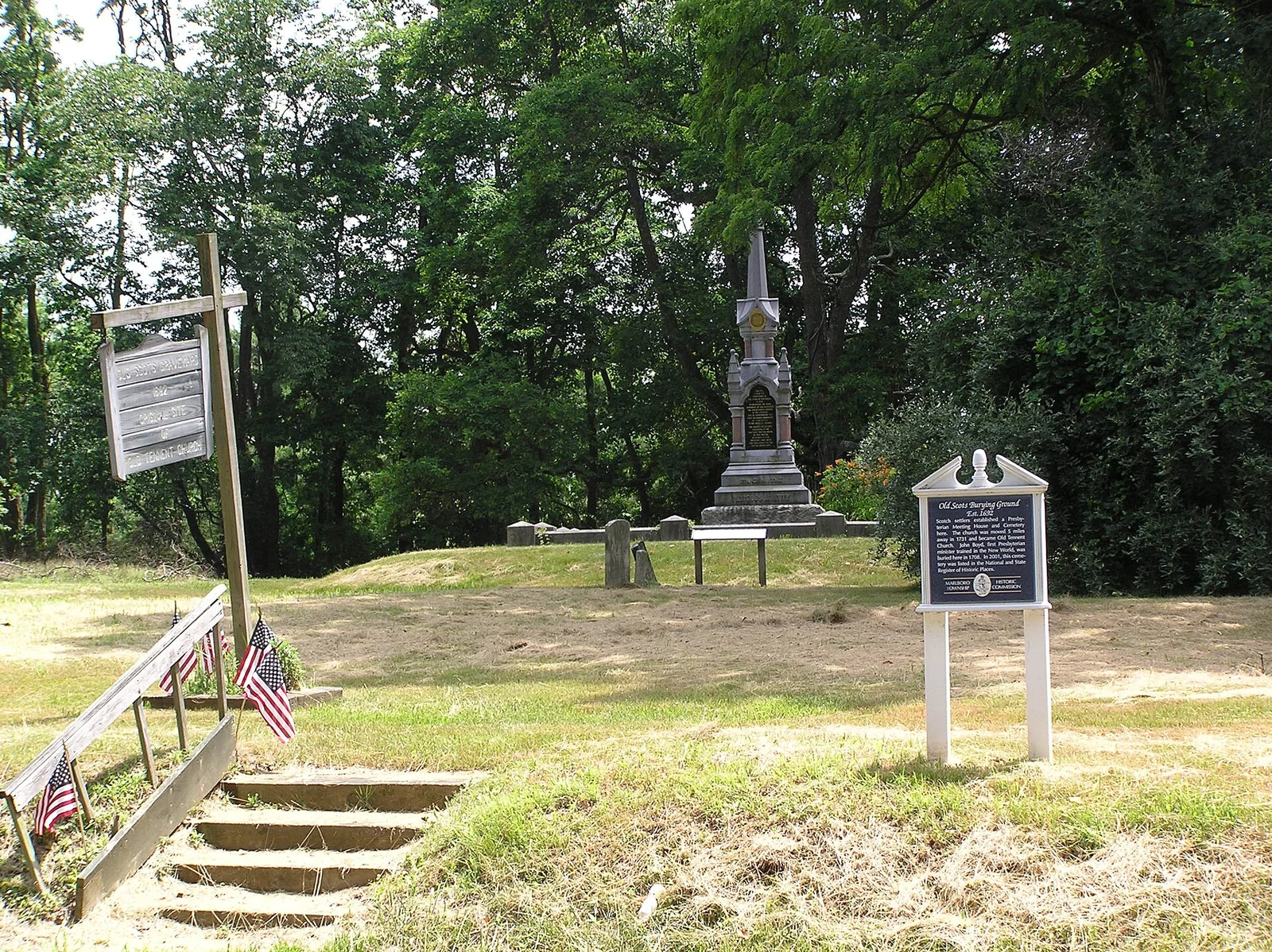 Grassy hilltop burial ground with an old stone monument, historic marker signs and small American flags by wooden steps