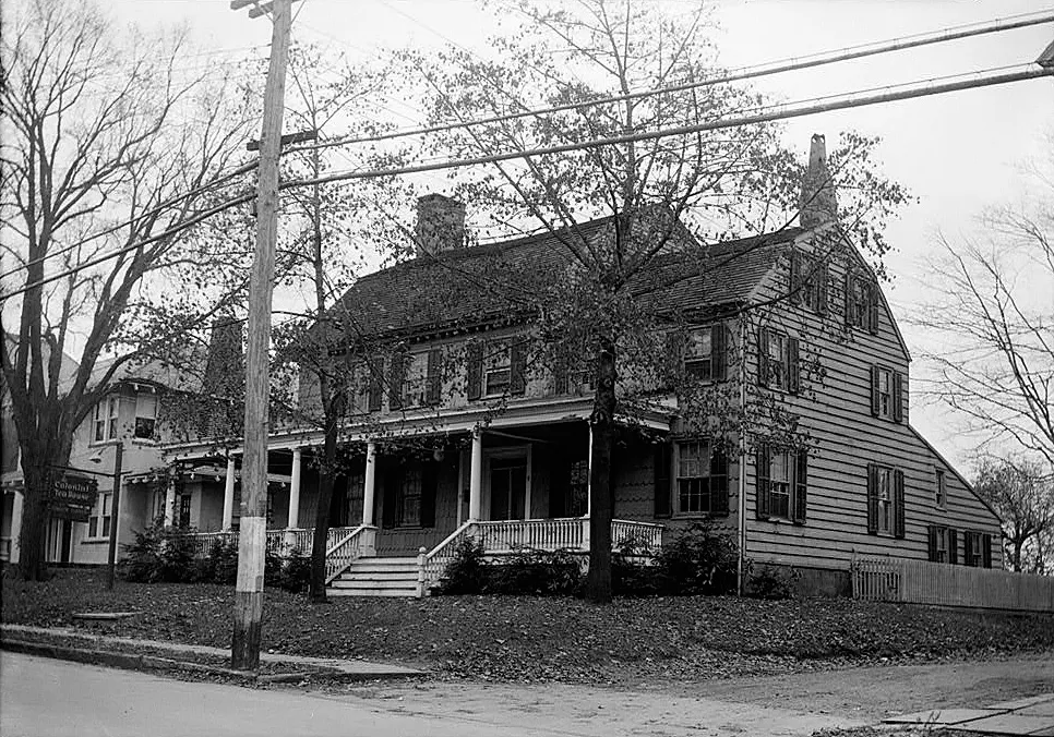 Black-and-white photo of a large colonial clapboard house with a long front porch behind bare trees