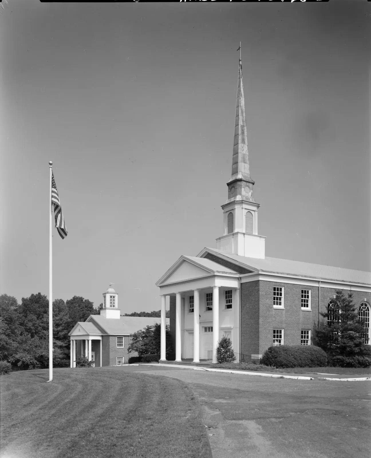 Black-and-white photo of a white-columned brick church with a tall steeple beside a flagpole on a wide lawn
