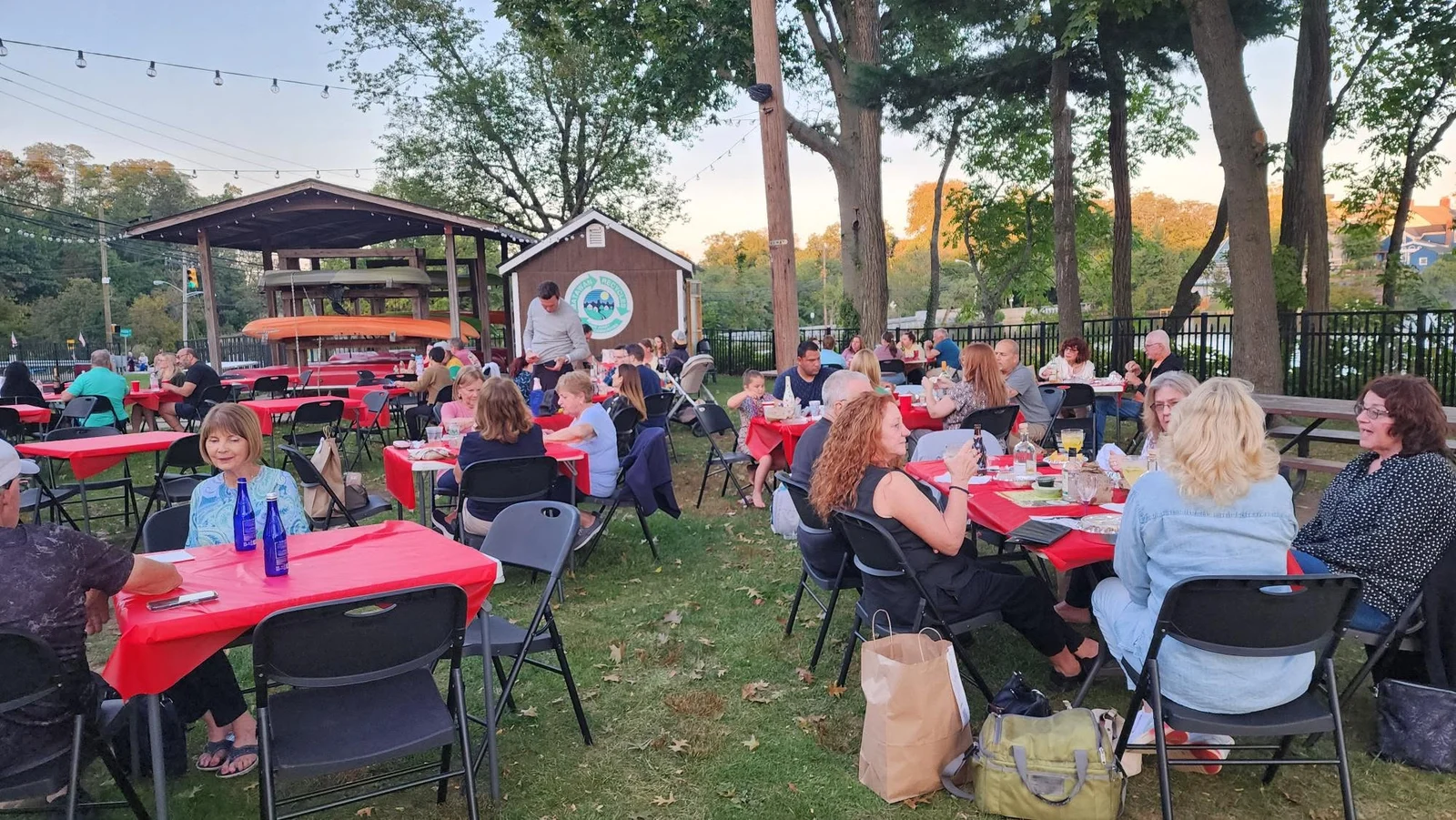Neighbors dining at red-covered tables on the lawn under string lights at dusk, beside a boathouse pavilion stacked with kayaks