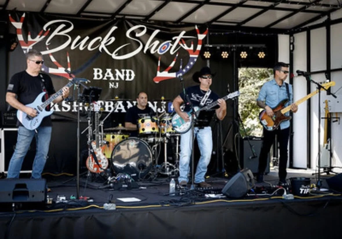 The Buck Shot Band playing guitars and drums on an outdoor stage under a stars-and-stripes banner