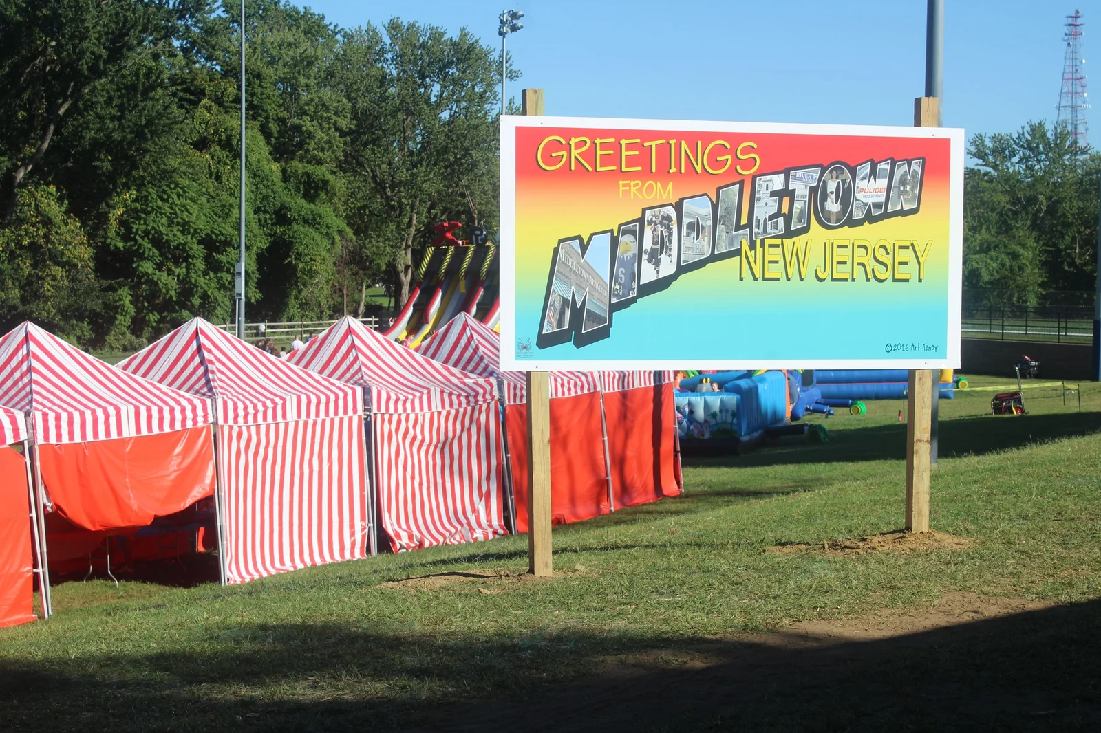 A colorful Greetings from Middletown, New Jersey sign on the lawn beside red and white striped carnival booths at Middletown Day