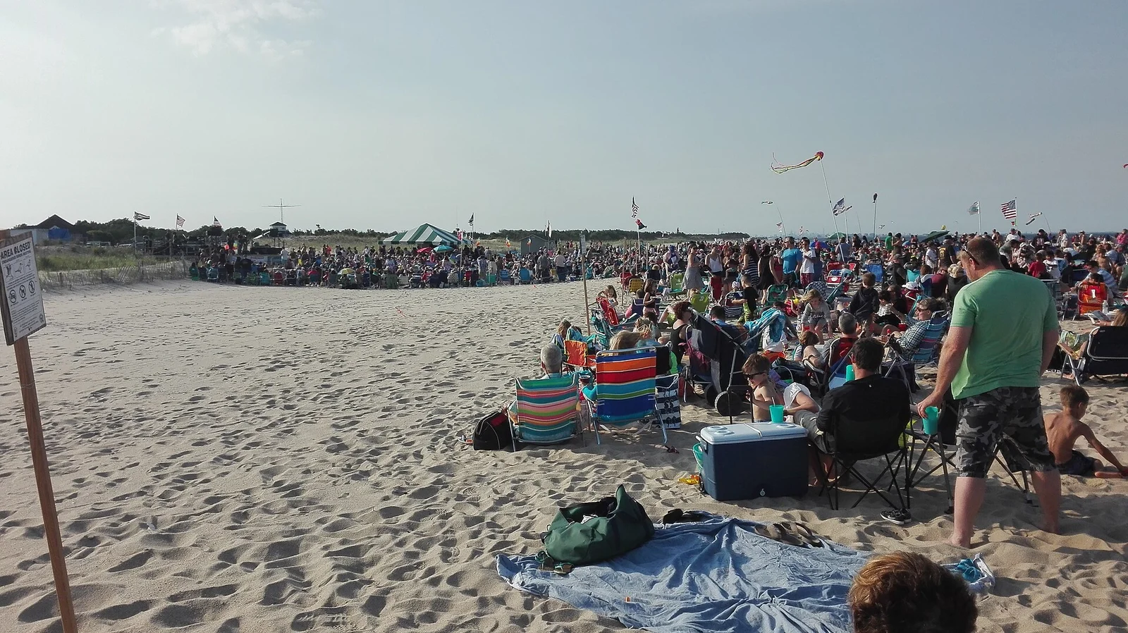 A large crowd in beach chairs and on blankets gathered on the sand at Sandy Hook for an evening beach concert, flags and a kite overhead