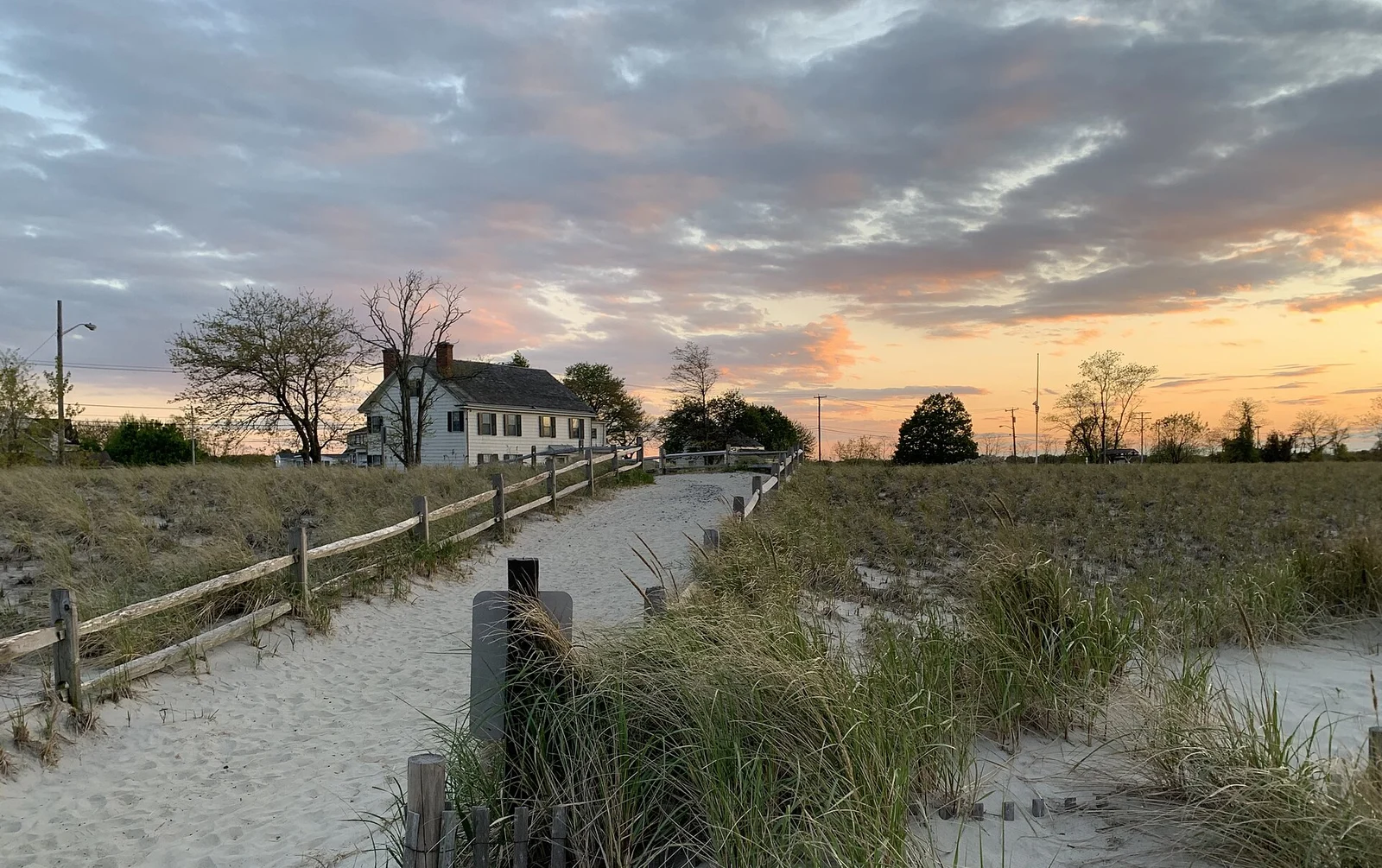 A sandy path through dune grass leading to the white Seabrook-Wilson House under an orange evening sky in Port Monmouth