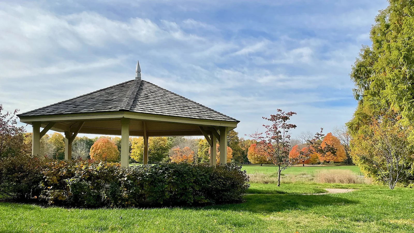A wooden gazebo on a green lawn with fall trees behind it at Thompson Park in Lincroft