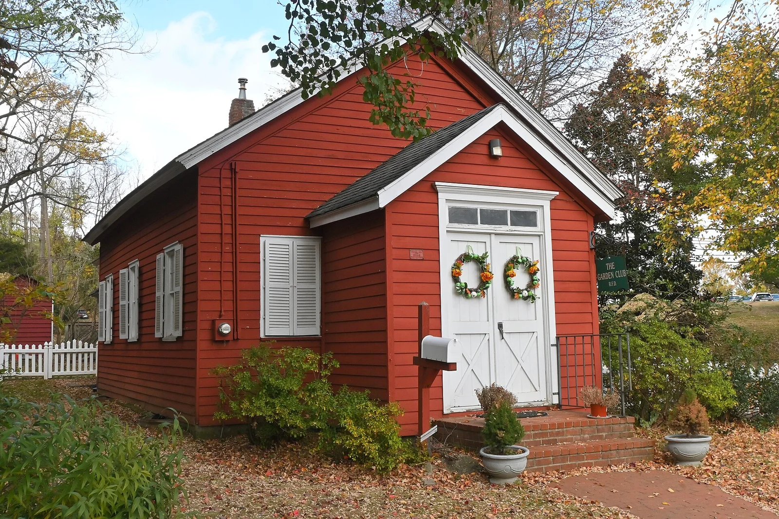 The small red Union Schoolhouse with white double doors decorated with wreaths, surrounded by autumn trees and potted plants