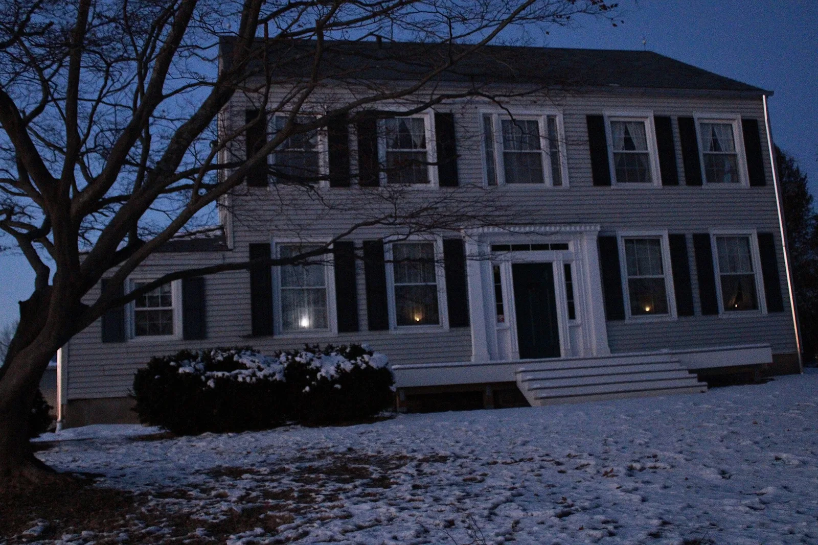 The two-story white Thomas Baird Homestead with black shutters, snow on the lawn at dusk