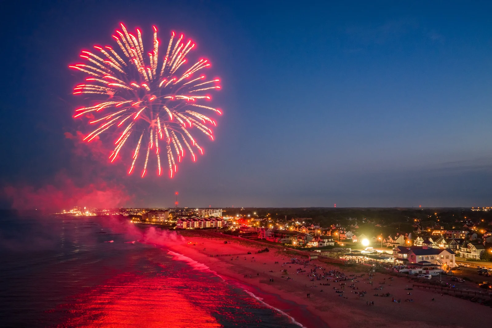 Red fireworks burst over the ocean at dusk, its glow reflected on the beach with the lights of the shoreline beyond