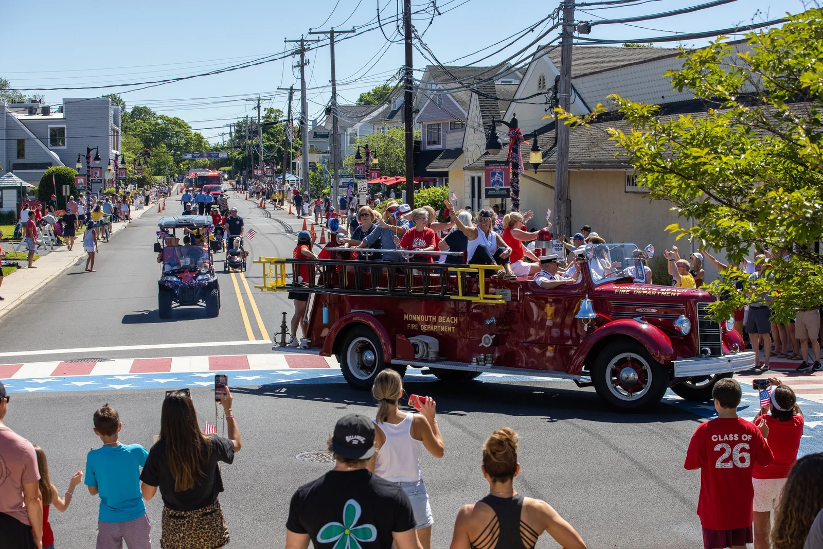 An antique red fire engine rolls down the parade route over a stars-and-stripes crosswalk as crowds wave from both sidewalks