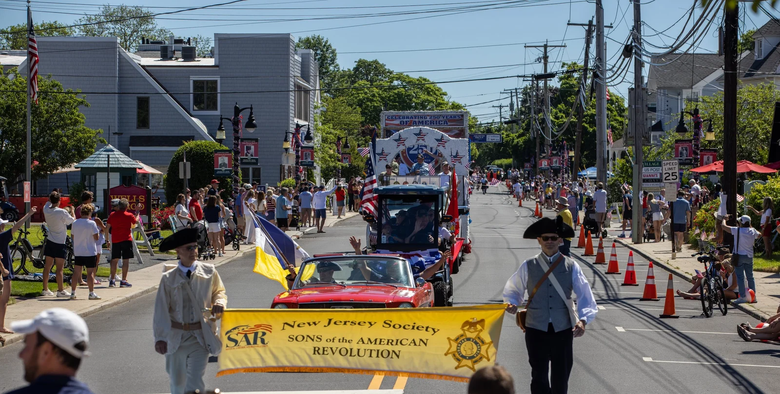 Marchers in colonial dress carry a Sons of the American Revolution banner ahead of a red convertible and floats, with crowds lining the street